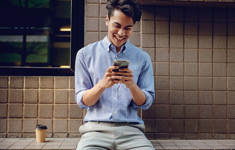 Man sitting against a tiled wall in a blue button-up shirt, smiling while looking at his mobile phone. Sitting to his left is a brown, disposable coffee cup.