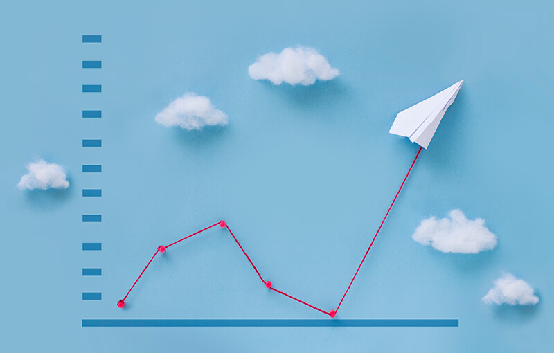A 3D line graph showing improvement against a light blue background. The line is made of red string and at the end of the string is a paper airplane. There are five small bits of cotton representing clouds sitting against the background, making it appear that the line and paper airplane are flying off into a sky.