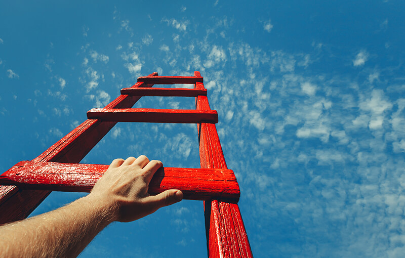 A wooden ladder painted red pointing up into a blue sky. A hand rests on the lowest rung of the ladder.