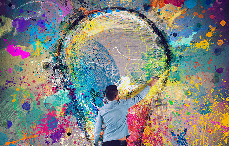 Man painting a large lightulb on the side of a wall. The wall is covered in colorful paint splatters.