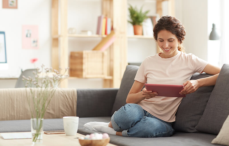 A smiling woman sitting on a couch and looking at a tablet with her legs folded up under her.