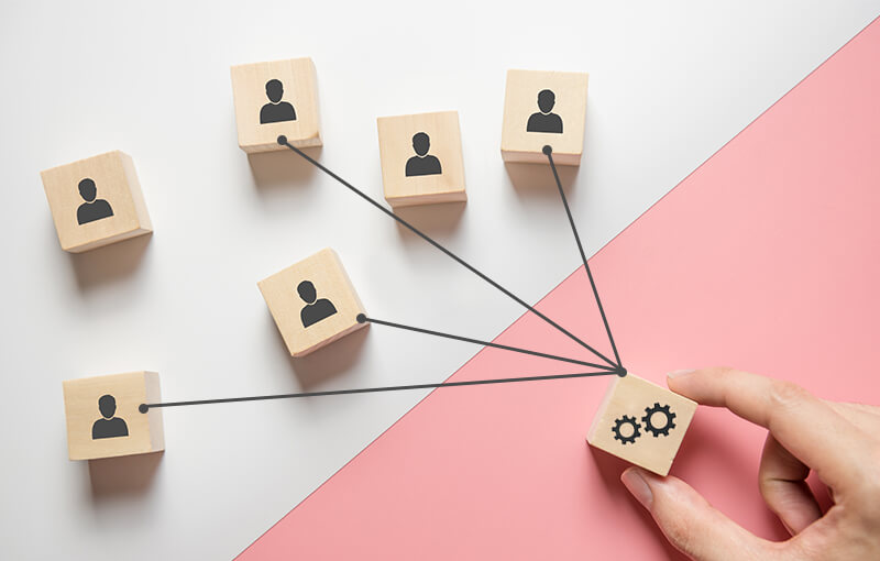 Wooden blocks resting on a white and pink background. Six of the blocks have icons of people; the remaining block has a set of gears. Four lines are drawn coming off of the gear block and connecting to four of the blocks with the icons of people.