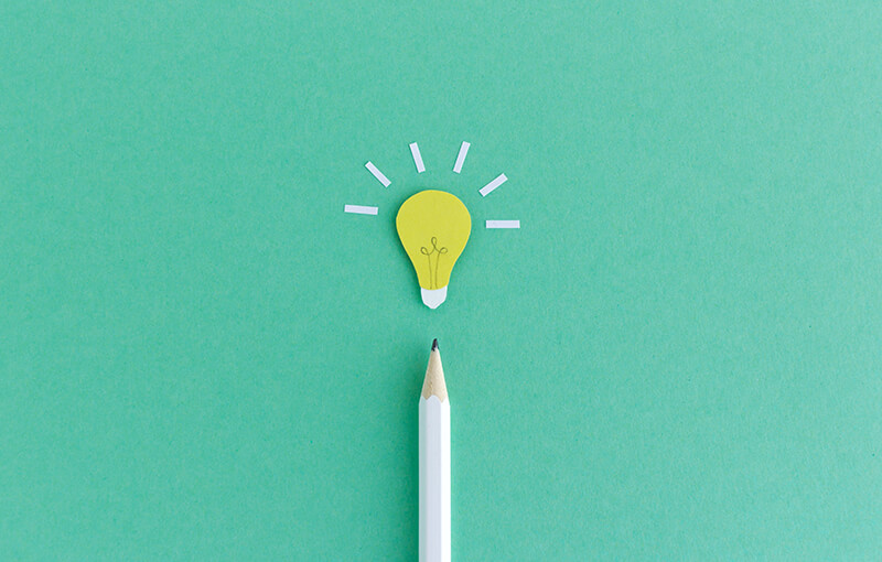 A white pencil, pointing to the top of the photo, resting on a seafoam green background. At the tip of the pencil is a paper light bulb cutout.