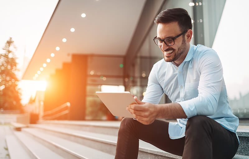 A person with short brown hair and glasses sits on the edge of some stairs in front of a large corporate building. They're smiling while they hold a tablet. - Neovation Learning Solutions