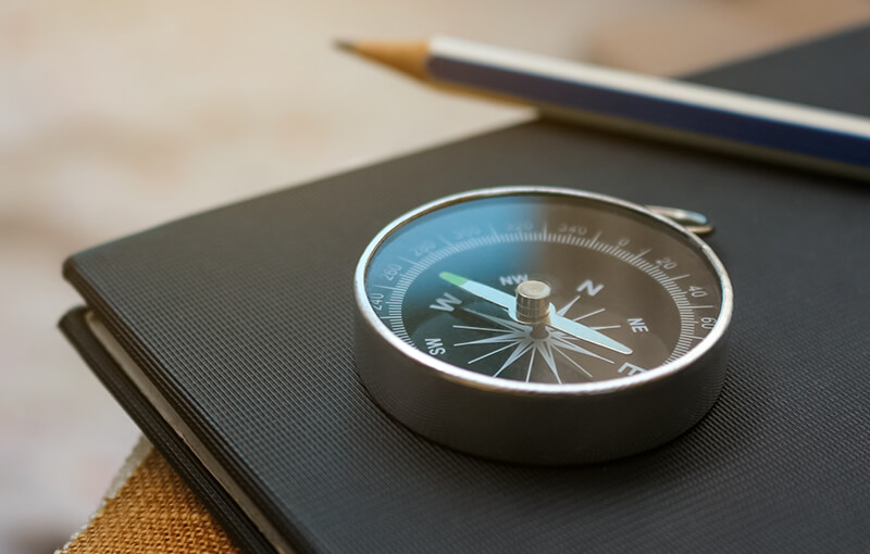 Compass resting on a black notebook with a pencil sitting beside it, slightly blurred in the background.