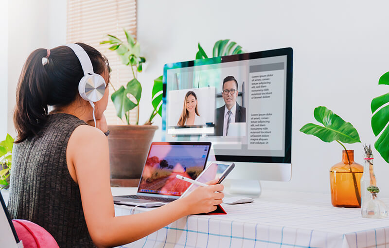 Young woman with long, dark hair pulled back in a high ponytail, wearing headphones. She sits at her desk in front of her laptop and monitor having a virtual call with colleagues, while also holding a tablet and pen.