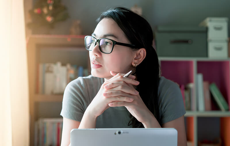Young woman looking to the left, out a window, a thoughtful expression on her face. Her hands are clasped together under her chin and her laptop sits in front of her.