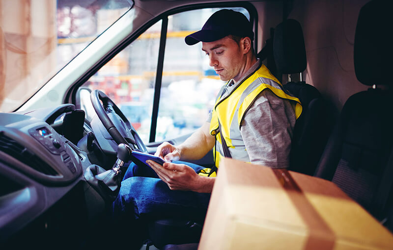 Delivery driver wearing a dark blue baseball cap and yellow safety vest, sitting in a parked car, looking at his digital tablet.
