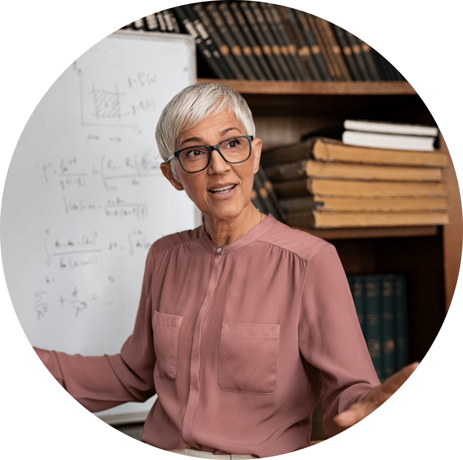 A white haired woman in a pink shirt stands in front of a whiteboard in a classroom.