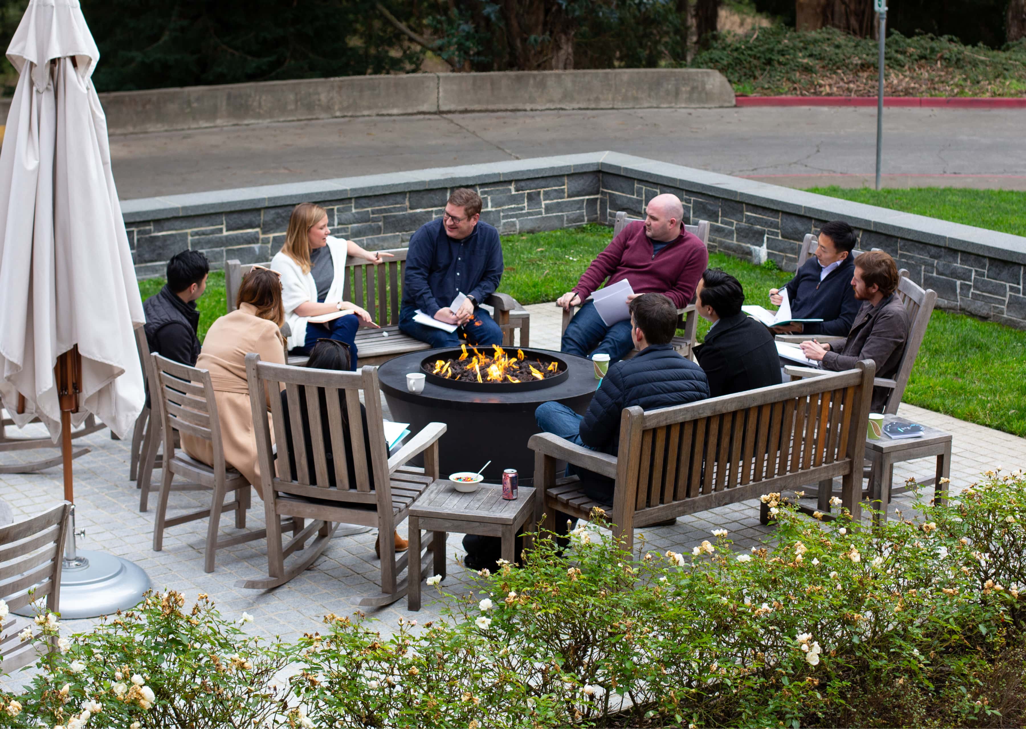A group of Praxis fellows talk together while seated around an outdoor firepit.