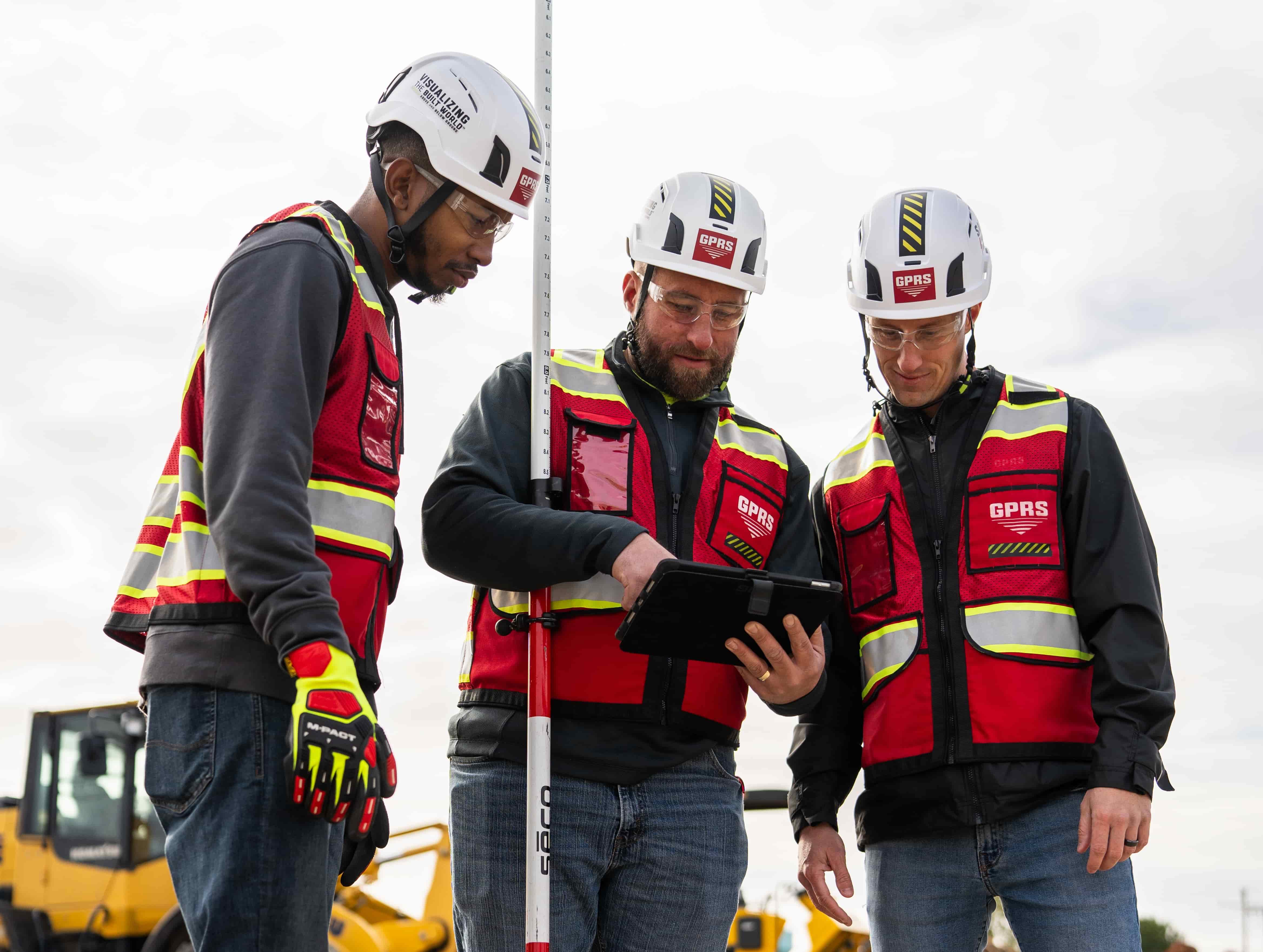 Three men in personal protective equipment look at a tablet.