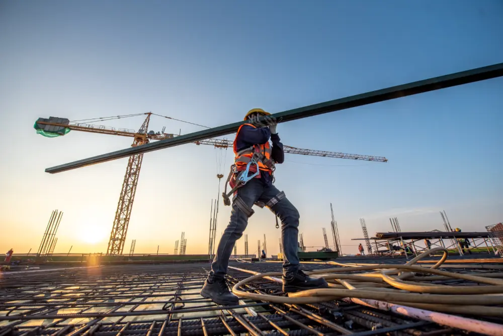A construction worker carries a long beam across a job site.