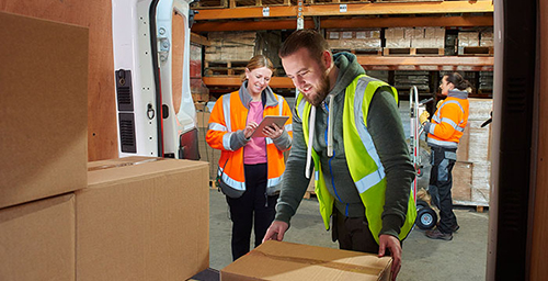 An image of one warehouse worker moving boxes off a truck and another warehouse worker using software on a tablet to track the inventory in the boxes as they come off of the truck for proper inventory control.