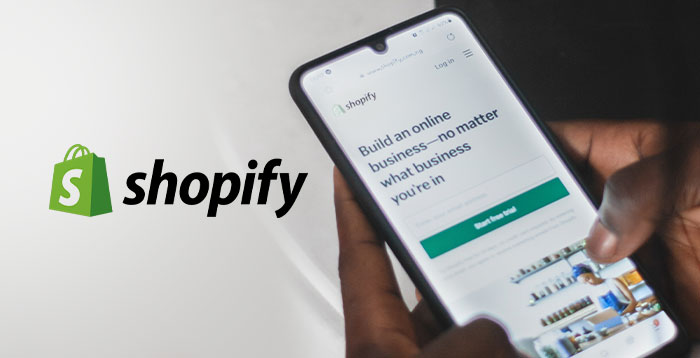 The Shopify logo shown on the left and the Shopify website is being used by a person on a mobile phone.