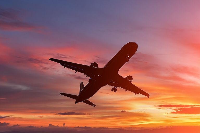 Silhouette of airplane flying across the sky as the sun is setting. The sky is lit up in shades of orange, yellow, pink and blue. 