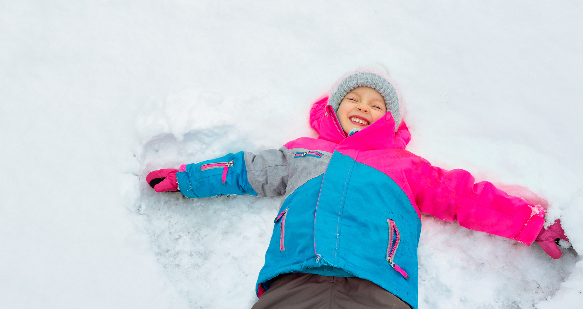 A young girl in a blue, grey and pink snowsuit makes a snow angel