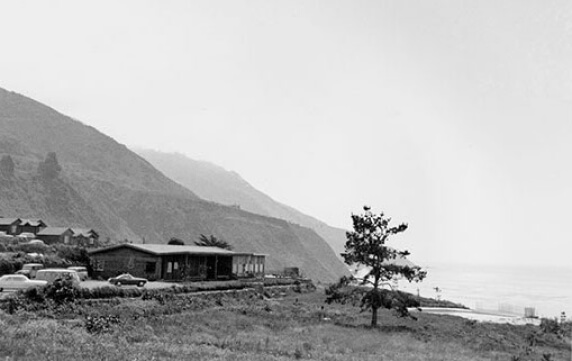 An black and white photo of a tree, a car, and a one-story building on a hill overlooking the ocean.