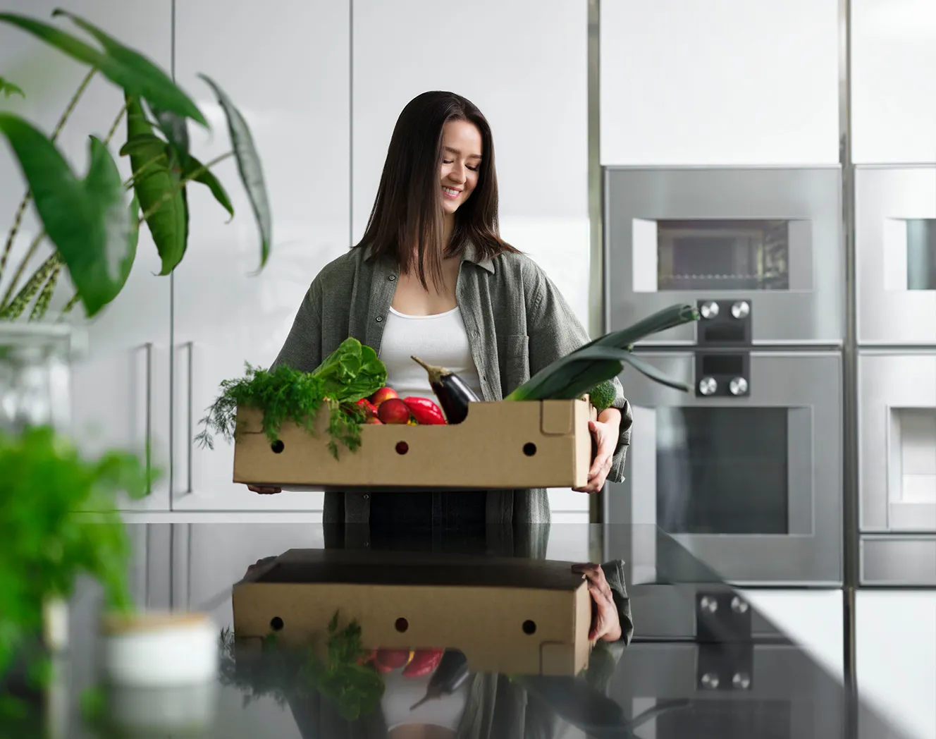 Woman holding a box filled with vegetables in a kitchen. Photo..