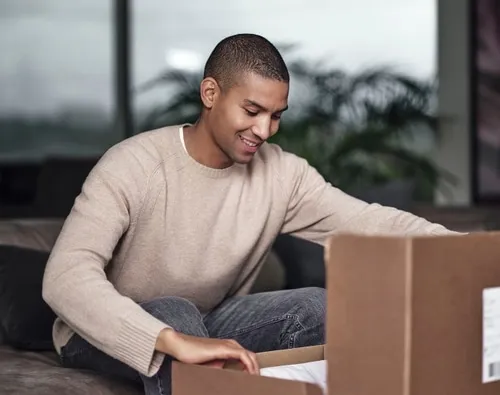 Young man sitting on a sofa, smiling, looking down and opening a cardboard box. Photo.