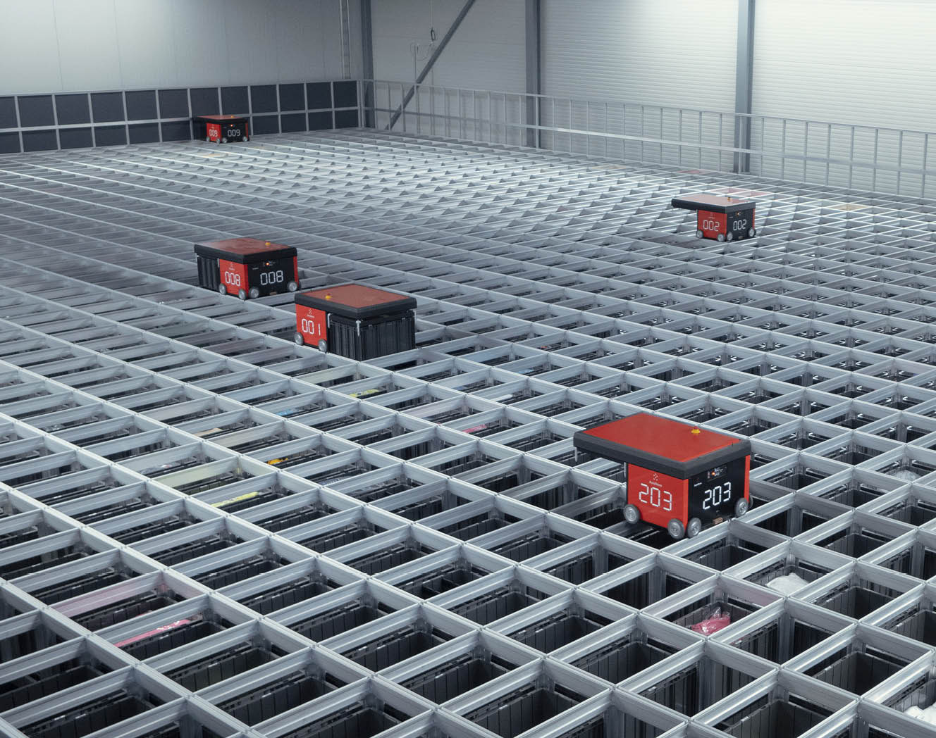 Blick von oben auf ein AutoStore Grid. Vier AutoStore Roboter des Typs R5 sind bei der Arbeit. Ein Roboter des Typs steht in der linken Ecke an einer Ladestation. Foto. 