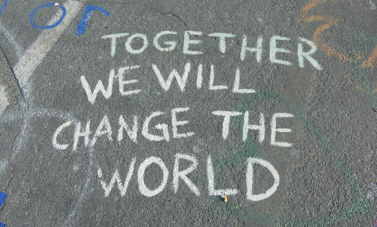 A piece of grey road with chalk marks on and the words 'Together we will change the world' written in capital letters using white chalk.
