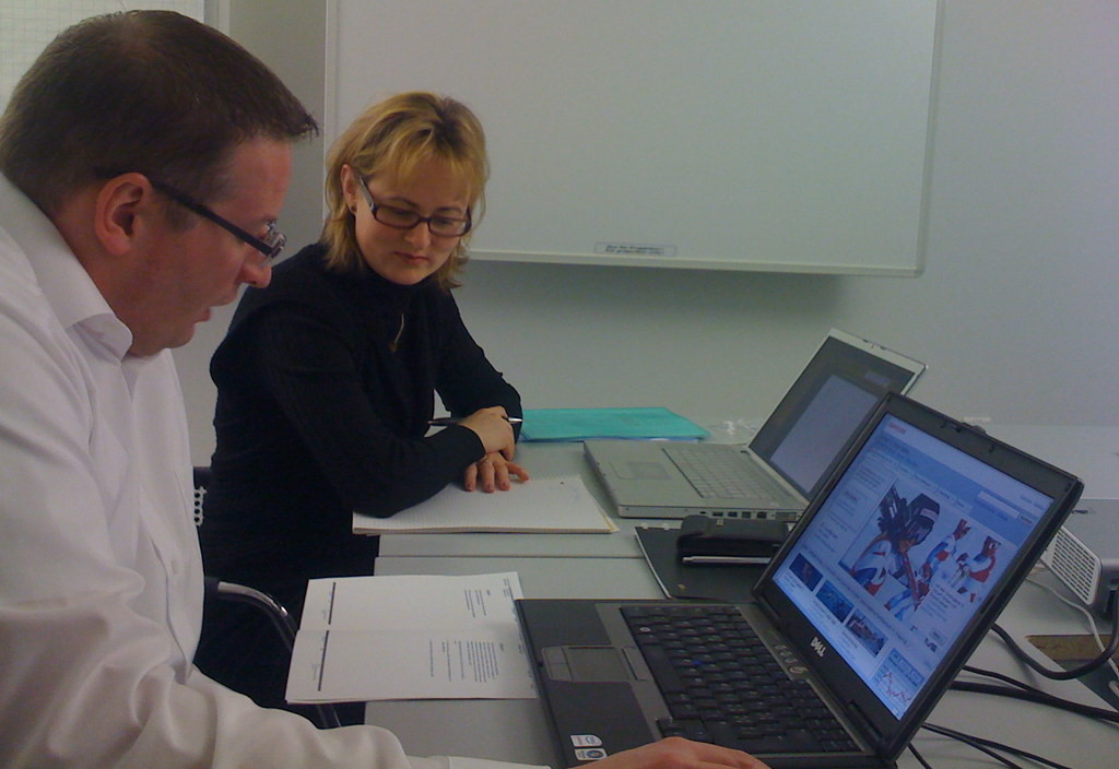 A man with dark hair and glasses reading a user testing task in front of a laptop while a woman with blonde hair and glasses looks on next to him.