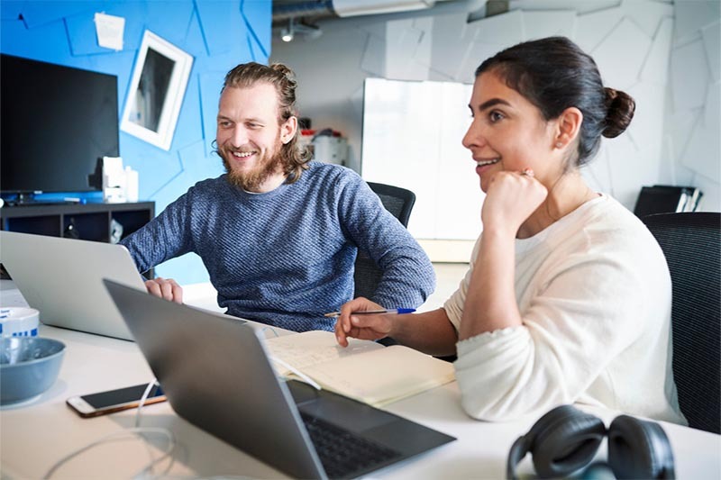 Zwei lächelnde Personen arbeiten im Büro an ihren Laptops