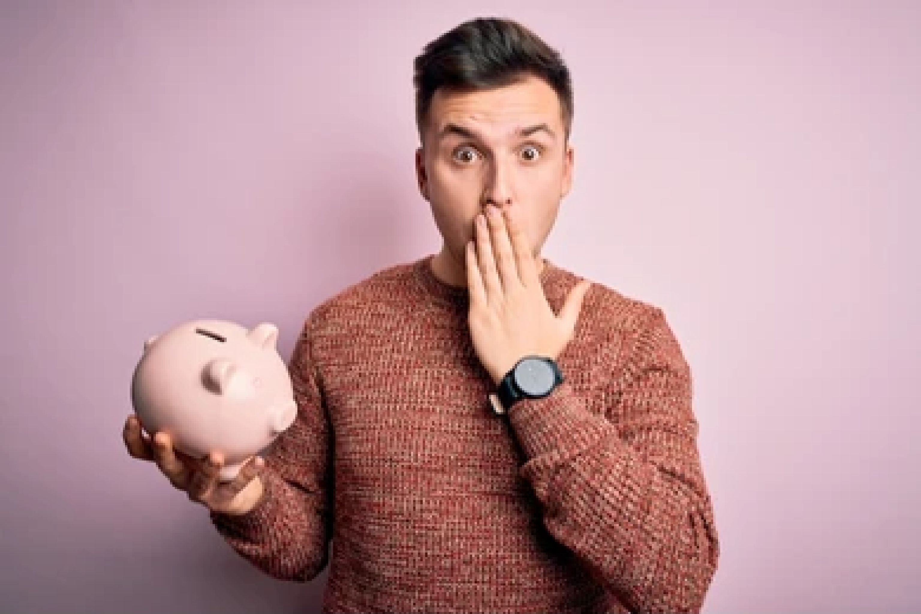 Young Caucasian man holding piggy bank for savings. Has his hand covering his mouth and his face appears shocked. 