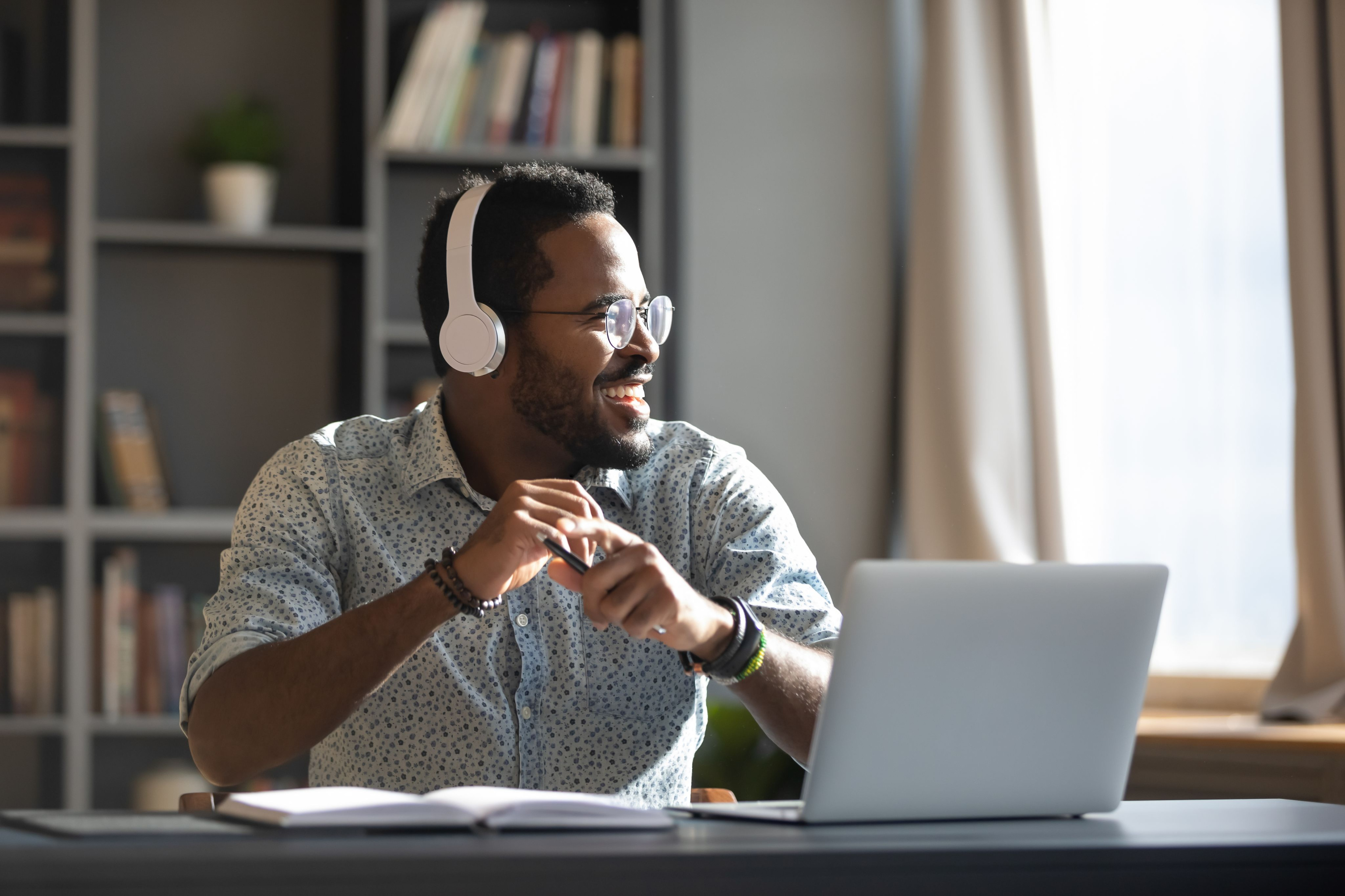 Happy relaxed man wearing wireless headphones, listening to a podcast. In front of him is a notebook and open laptop. He holds a pen in his hand.