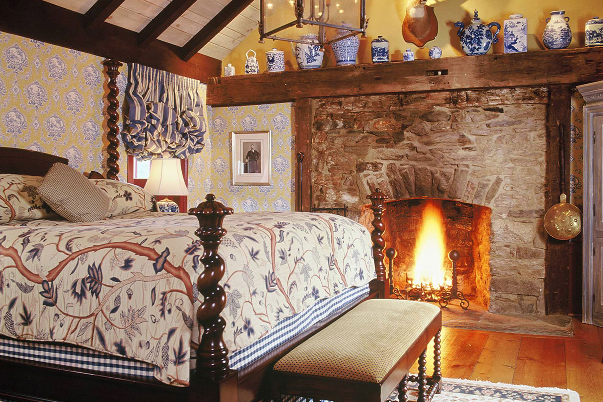 A large master bedroom featuring hardwood floors, a king sized bed and large stone fireplace with a blazing fire inside.