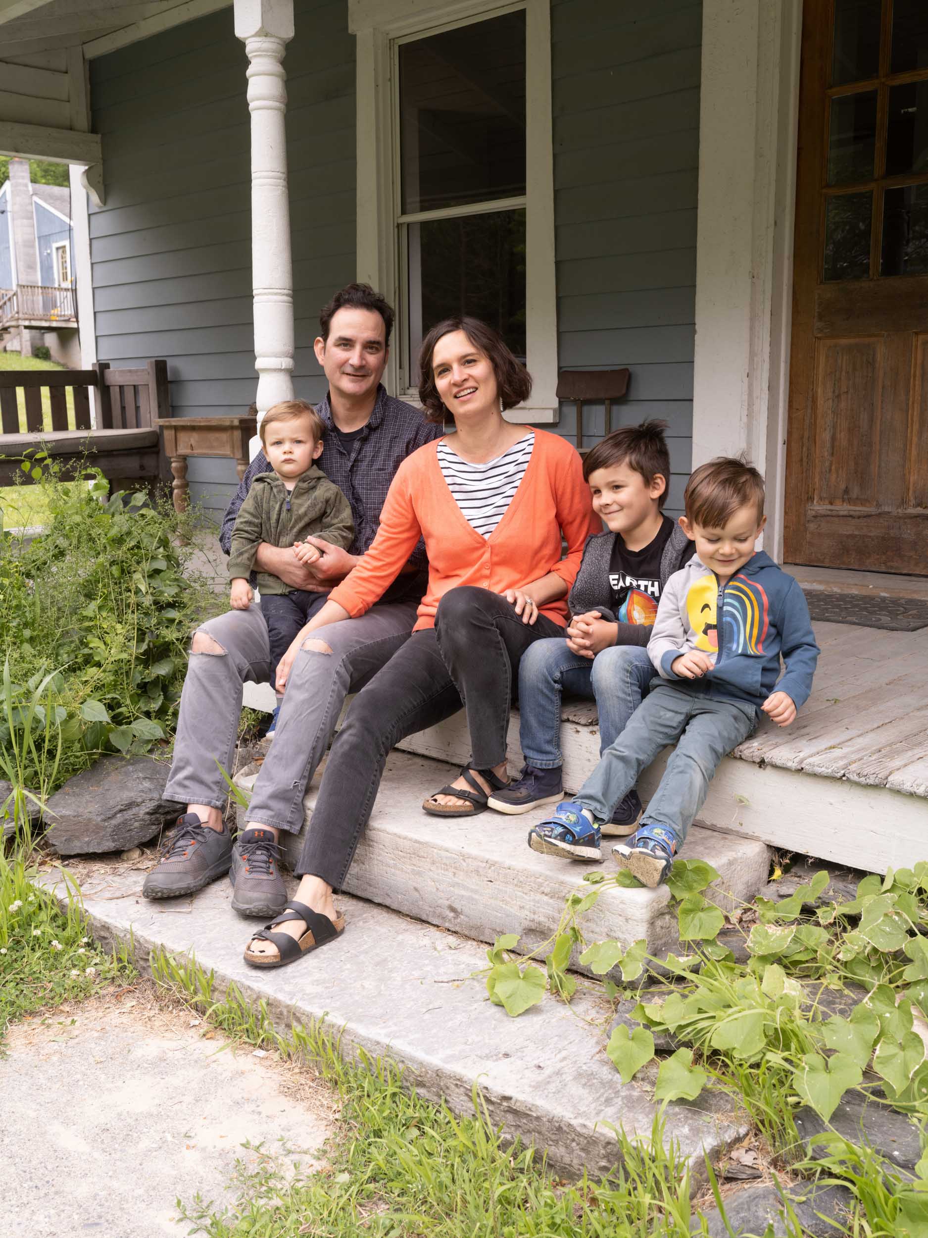 A man and a woman in a bright orange sweater sit smiling on a porch with their three children.