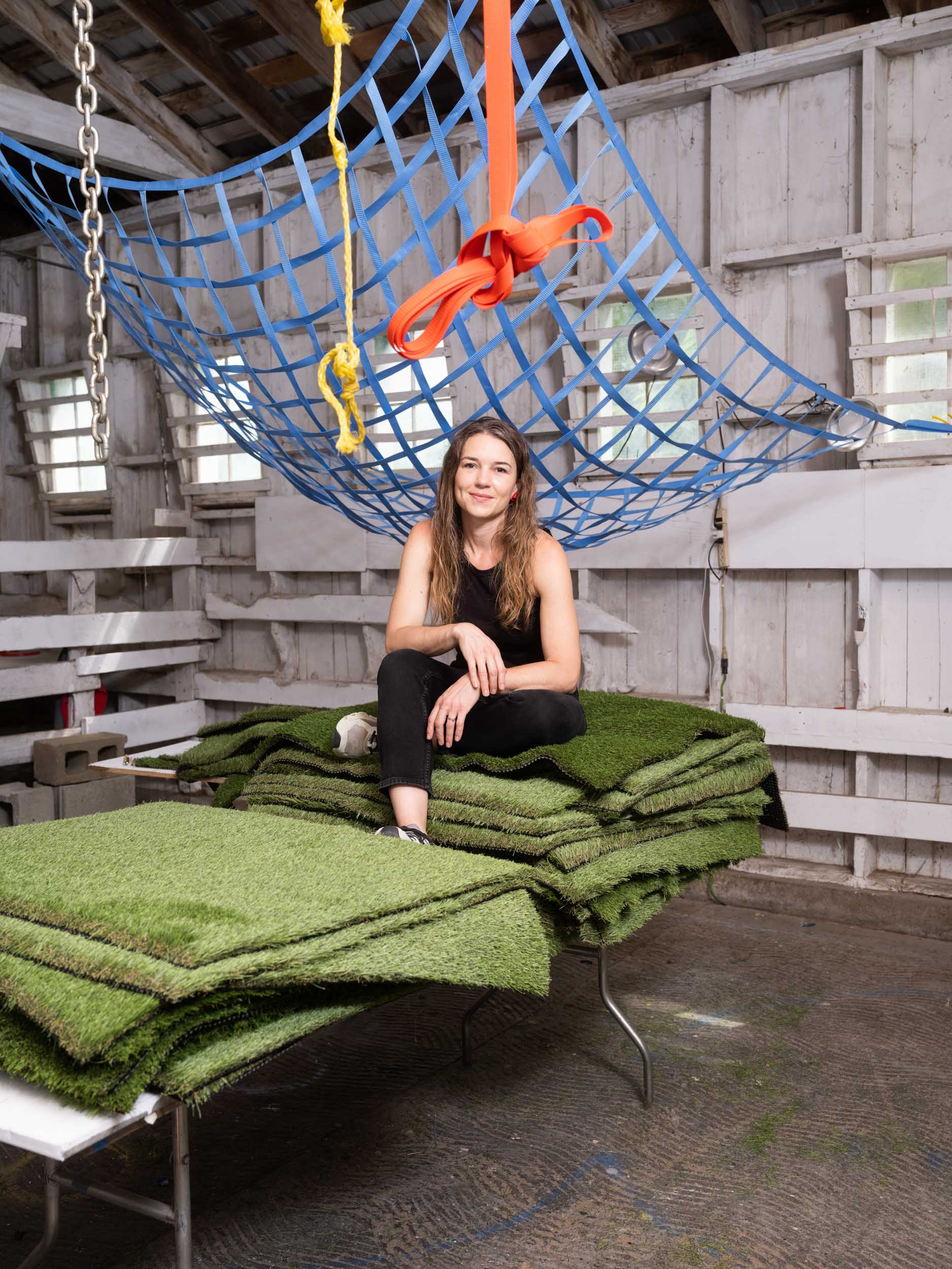 A woman sitting smiling on top of a pile of Astroturf on a folding table in a white barn studio. A blue net with orange ties hangs above her head.