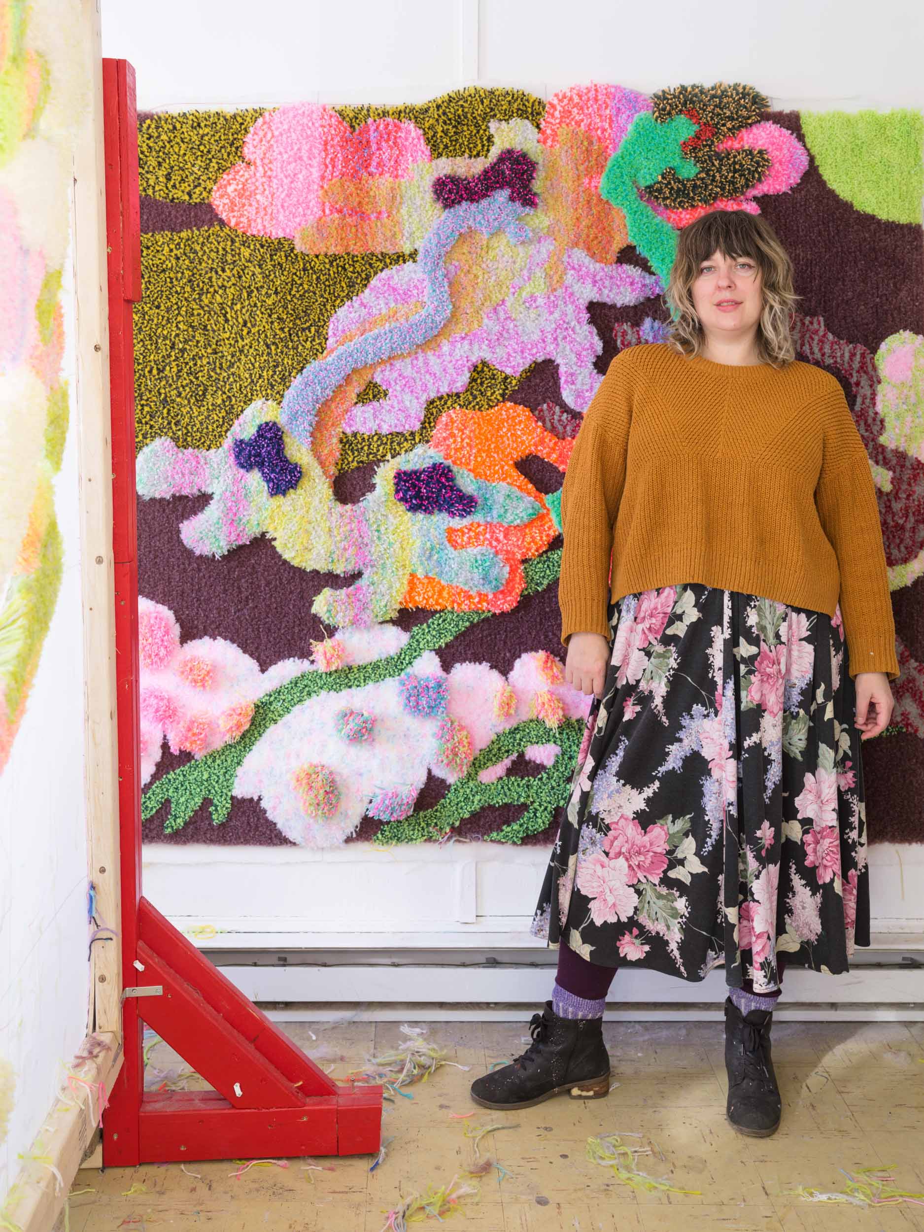 A woman stands in front of a large, colorful art rug hung on the wall. A red rugmaking frame outlines the left side of the photo.