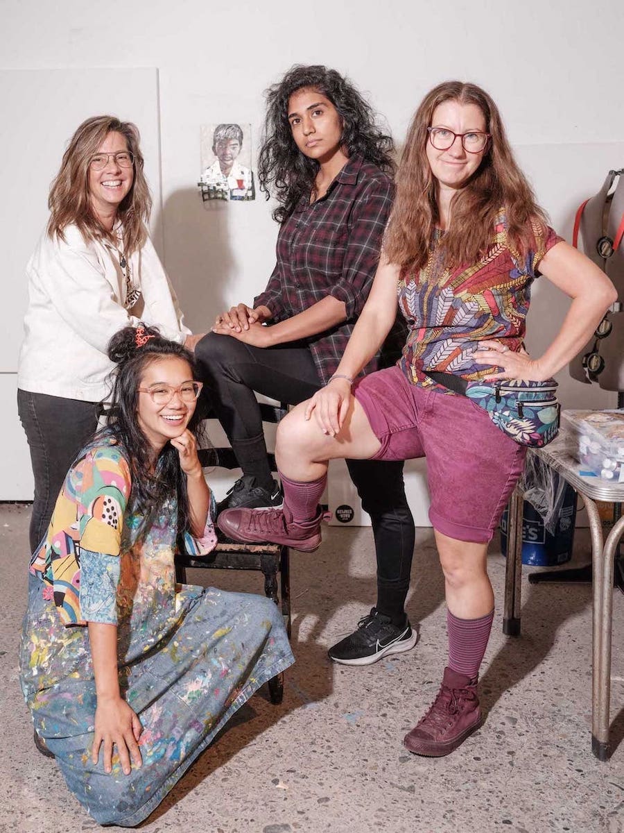 Four women smiling in an art studio. Three have one foot up on a chair, one is crouched in front.