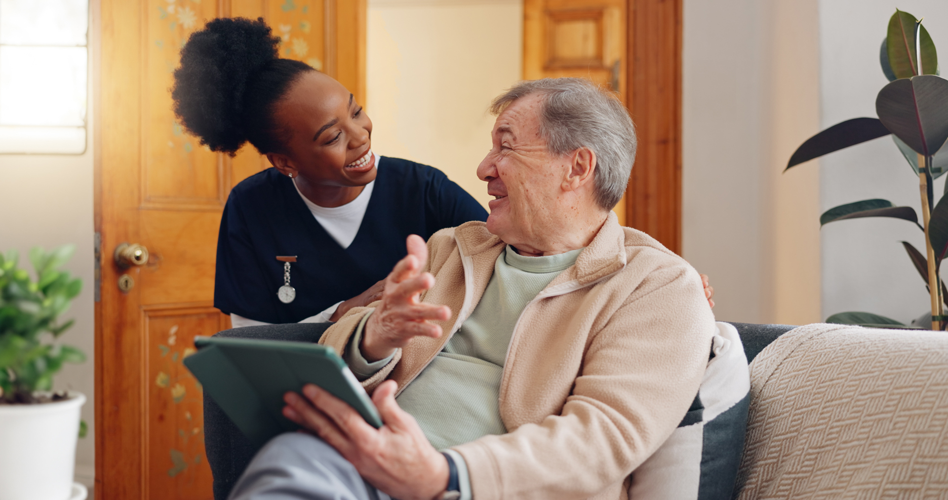 A female nurse checking in on their male patient who is holding a tablet inside of their home. 