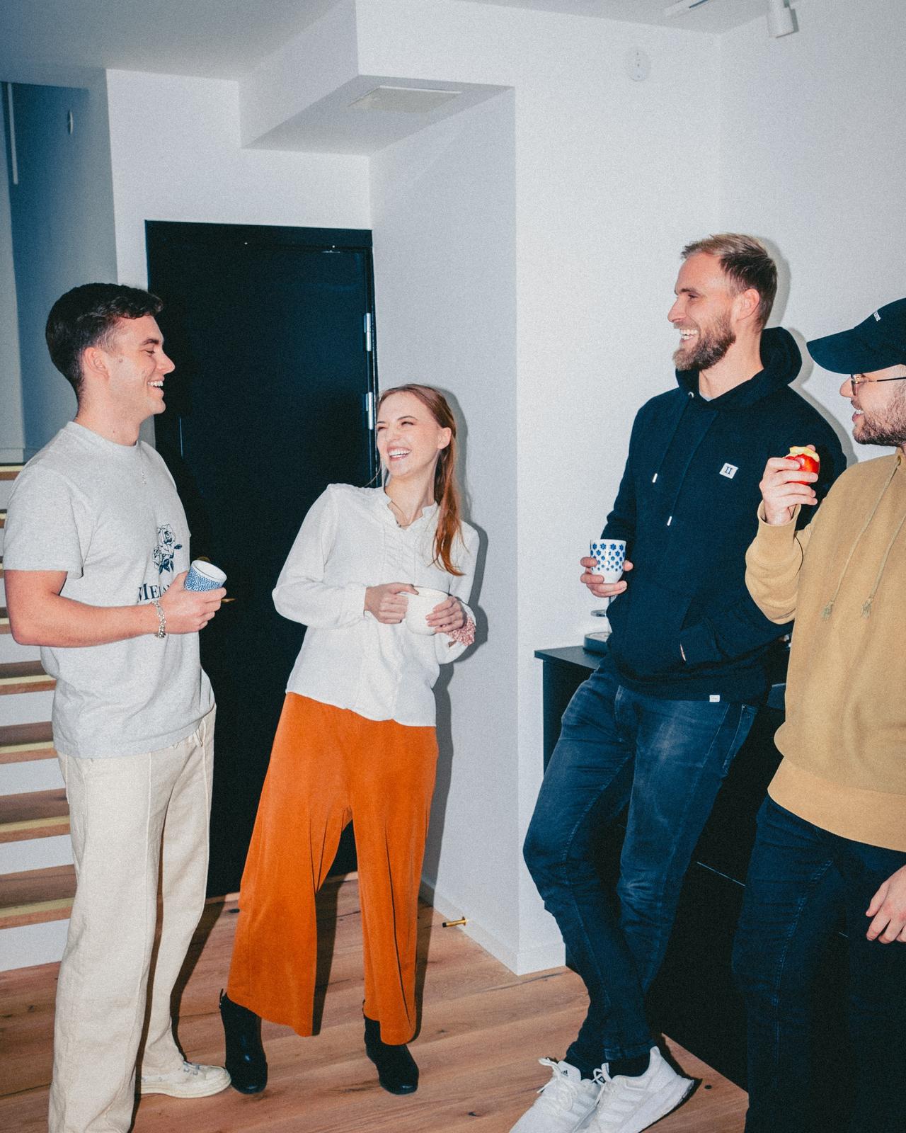 An edgy, high-contrast flash photo of four employees standing in a bright Scandinavian office kitchen together holding coffee mugs while genuinely laughing and smiling. The overall color of the picture is quite neutral, except for the female employee's pants, which are a saturated burnt orange