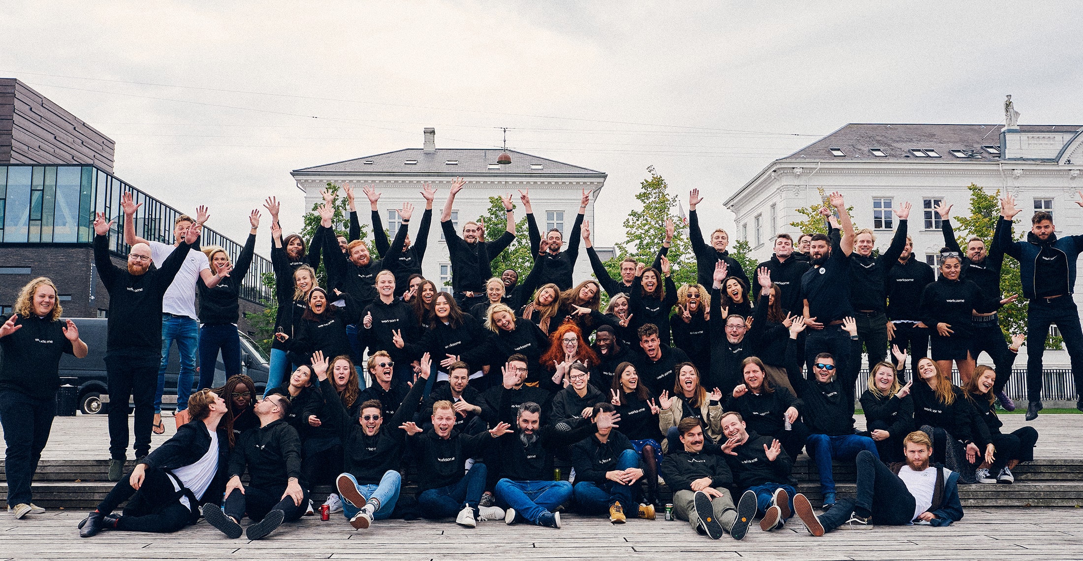 About 60 Worksome team members posing for a group photo outside with silly expressions and hands in the air. Everyone is wearing a black sweatshirt with the Worksome logotype.