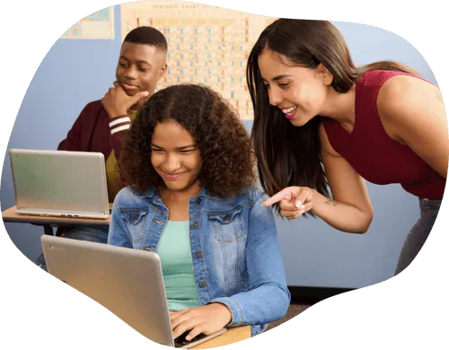 Two cheerful students are working on their laptops, while a teacher is talking to one of them and pointing at their laptop screen. 