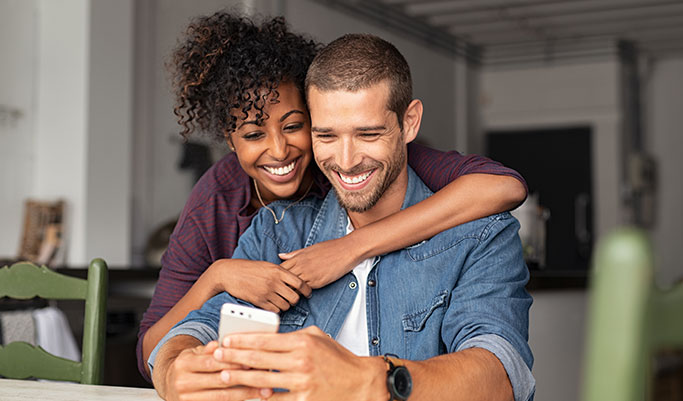 Man sitting with a woman wrapping her arms around him from behind, both looking at his mobile phone