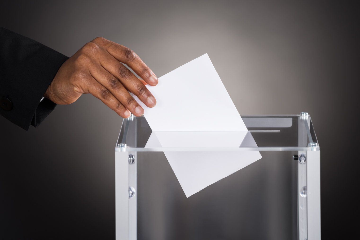 A person placing a ballot into a clear collection box.