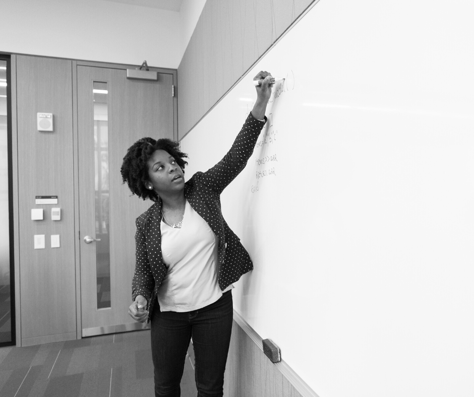 Image of a Black person with medium length hair wearing a dark blazer, white T-shirt, and a dark pants writing on a blackboard with their left hand