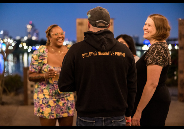 Three people talk and laugh against a night skyline back drop. 