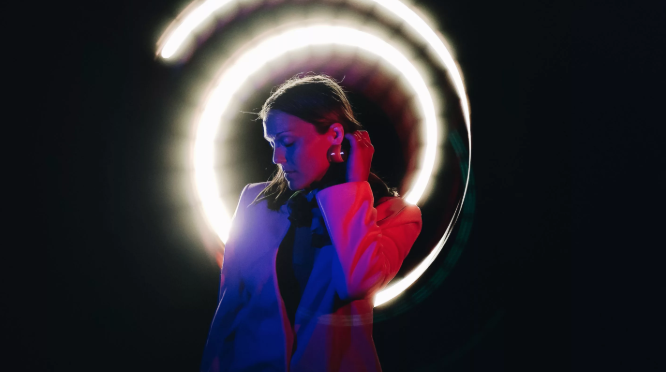 A woman in front of a black background, lit with pink and blue neon lighting