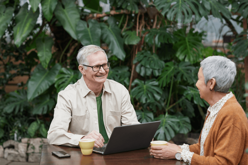 an older man with a laptop smiling sitting at a table with an older woman