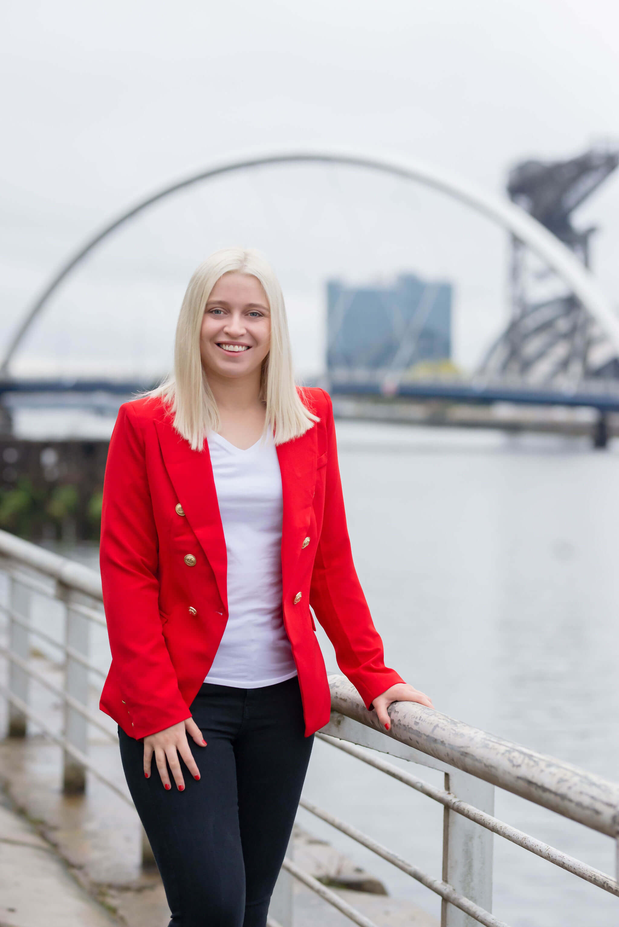 Emma is a young women in her 20s, wearing a bright red blazer standing along the Clyde River in Glasgow, with the Squinty Bridge in the background