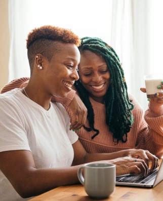 A female typing on her laptop with her female lover leaning on her shoulder
