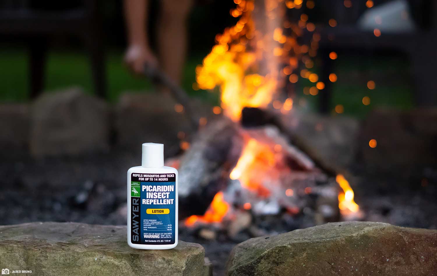 Insect repellent bottle sits in on rock in front of fire outdoors