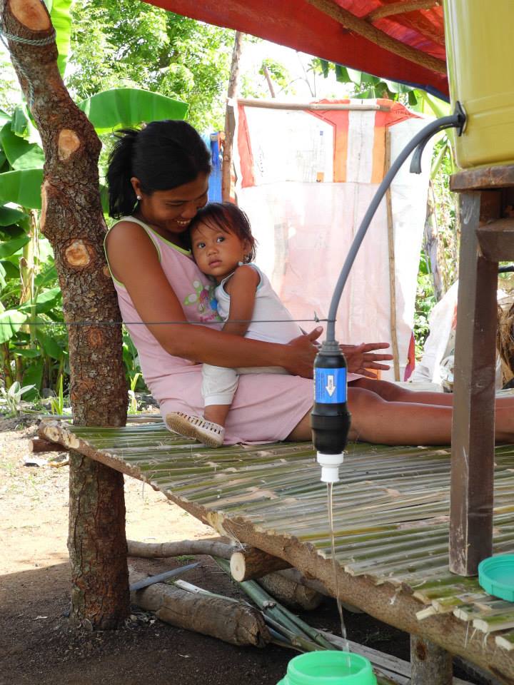 A Woman Holding Her Child in the Background while a Sawyer Water Filter Cleans Water