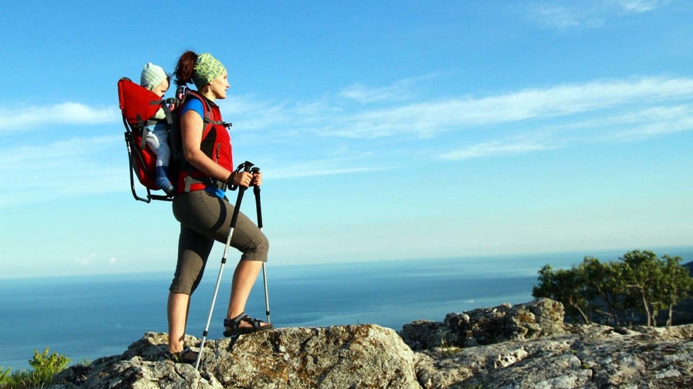 Woman with child in backpack carrier with trekking poles on a rock above ocean