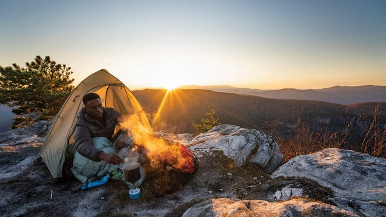 Hiker sitting in door of tent making food with camp stove